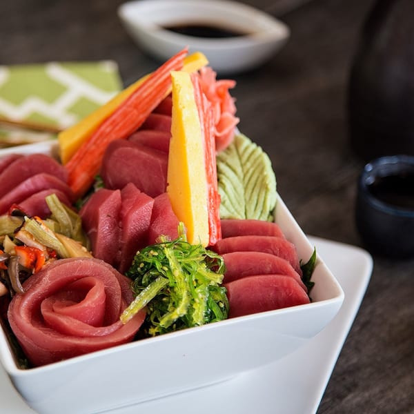A white bowl filled with various sashimi, including tuna, crab sticks, seaweed salad, and pickled ginger.