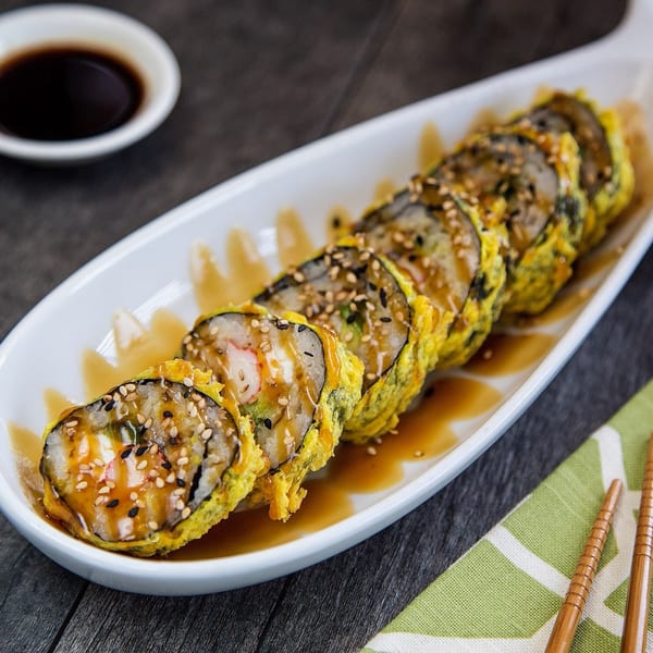 Tempura sushi rolls with sauce and sesame seeds on a white platter, with soy sauce and chopsticks.