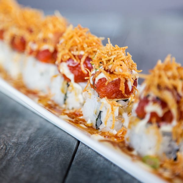 A long white plate of sushi rolls topped with red tuna, white sauce, and crispy fried onions.