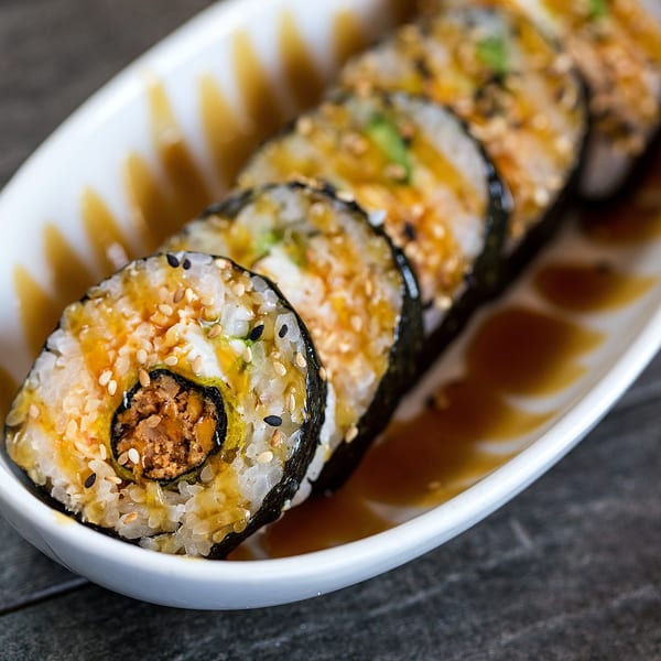 Close-up of sushi rolls with sesame seeds and sauce on a white rectangular plate.