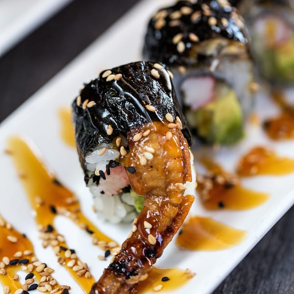 Sushi rolls with eel, avocado, and crab, drizzled with sauce and sesame seeds on a white plate.
