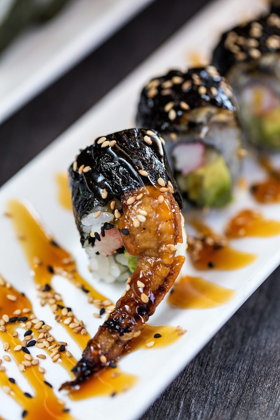 Sushi rolls with eel, avocado, and crab, drizzled with sauce and sesame seeds on a white plate.