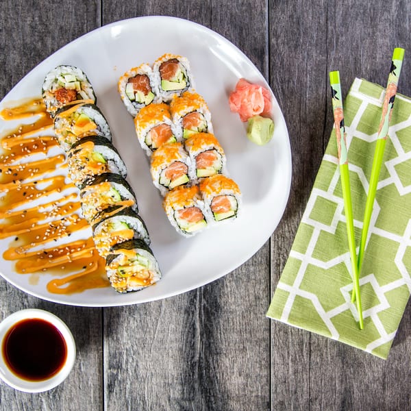 Two rows of sushi rolls with sauces, ginger, wasabi, soy sauce, and chopsticks on a wooden table.