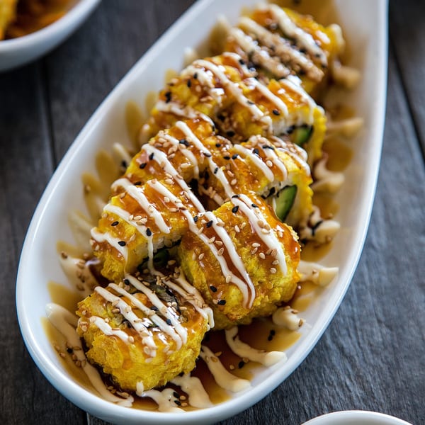 Golden fried sushi rolls drizzled with white and brown sauces, topped with sesame seeds on a white platter.