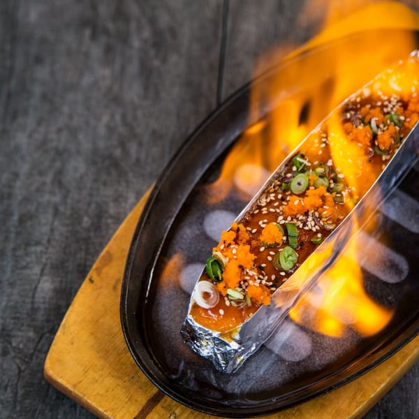 Flaming sushi roll in foil, topped with roe, sesame seeds, and green onions, on a sizzling plate.