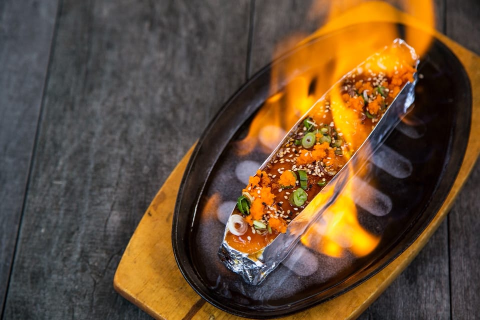 Flaming sushi roll in foil, topped with roe, sesame seeds, and green onions, on a sizzling plate.