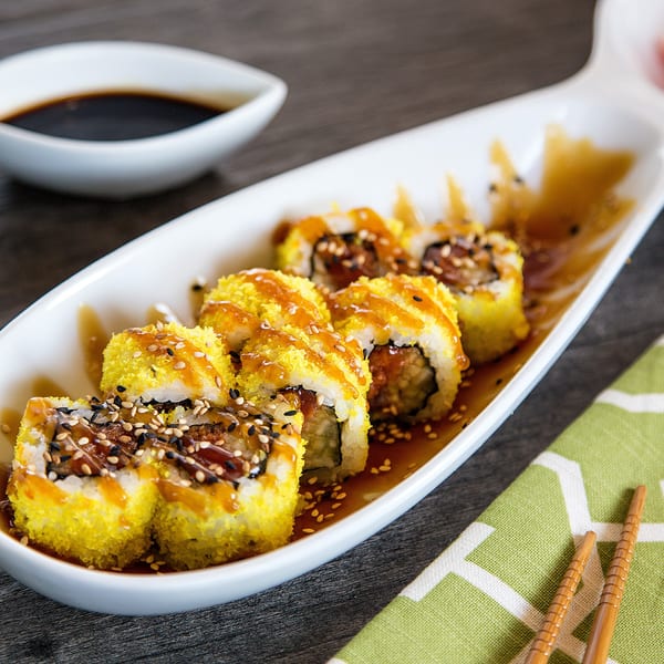 Sushi rolls with yellow topping and sauce on a white plate, with soy sauce and chopsticks.