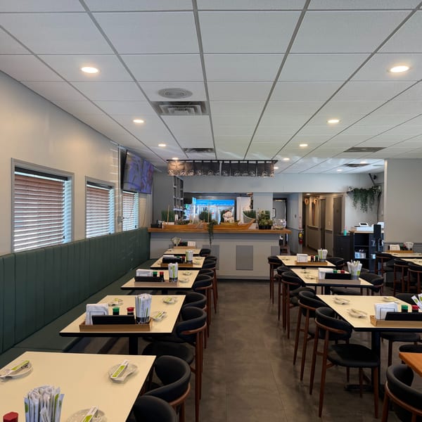 Empty restaurant dining room with green booth seating, light tables, and dark chairs, leading to a bar area.