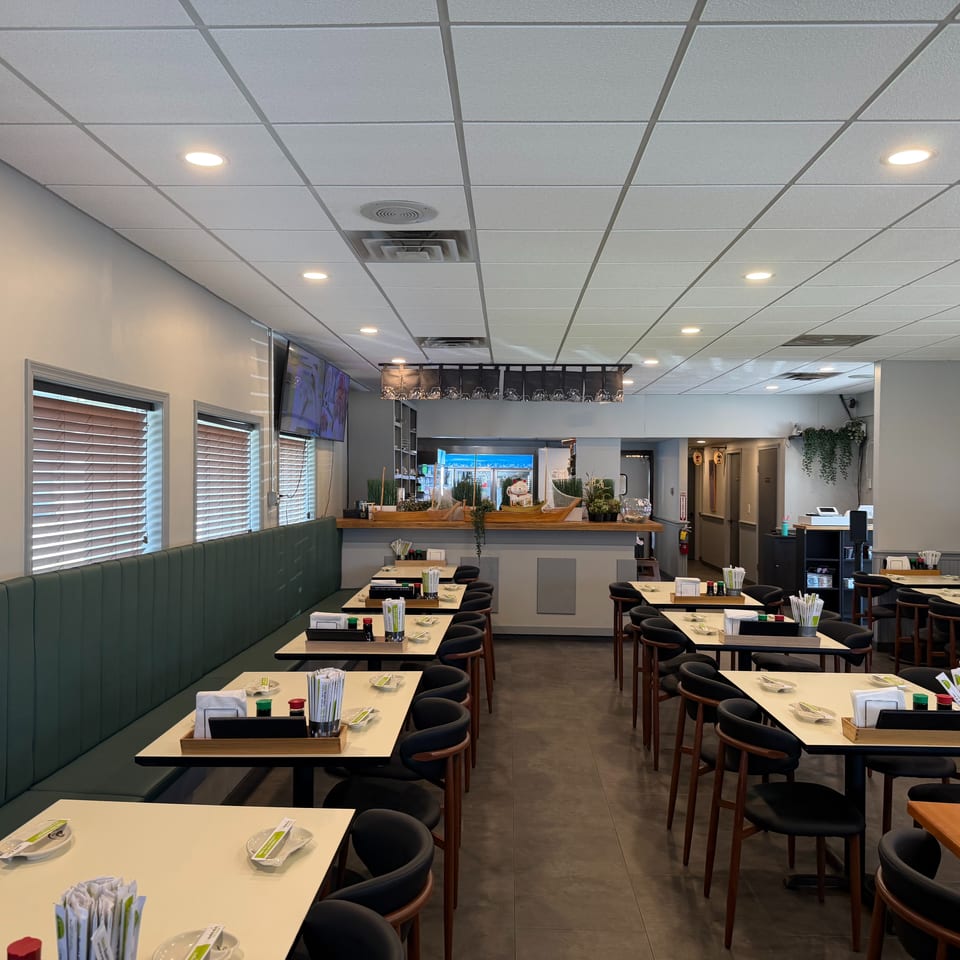 Empty restaurant dining room with green booth seating, light tables, and dark chairs, leading to a bar area.
